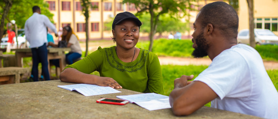 Two people sitting in a park speaking to one another and working from notebooks