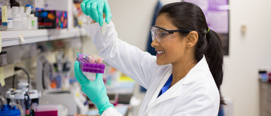 A scientist wearing a lab coat holds up and views a test sample