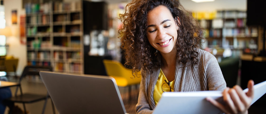 A student sits in a library reading a book while working at a laptop